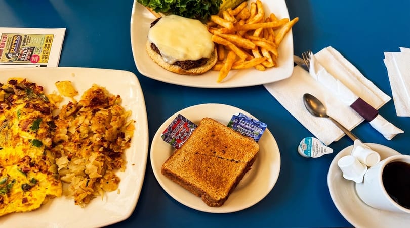 A diner table with a cheeseburger and fries, an omelet with hash browns, toast, and coffee.