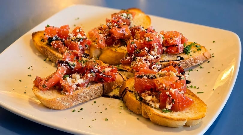 A white plate holds several pieces of bruschetta topped with diced tomatoes, cheese, and balsamic glaze.