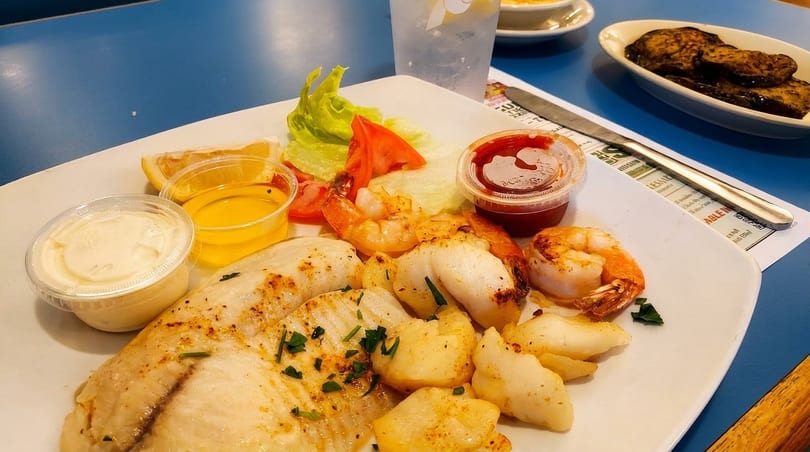Grilled fish, shrimp, and scallops with lemon, lettuce, tomato, and dipping sauces on a white plate.