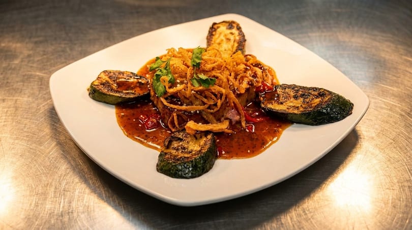 A gourmet dish featuring steak topped with crispy fried onions, surrounded by grilled zucchini and a rich sauce.
